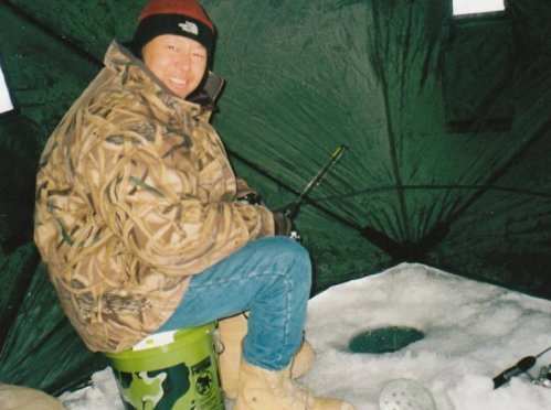 A person sitting on a bucket inside an ice fishing tent, wearing a camouflage jacket and winter boots, with fishing gear nearby.