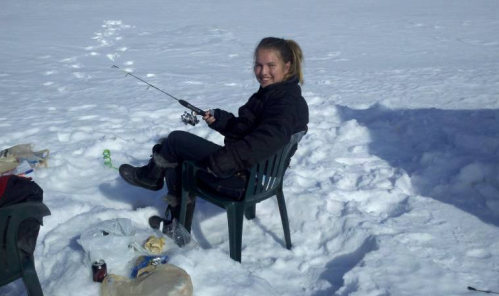 A person sitting on a chair in the snow, fishing with a rod, surrounded by ice and a few items on the ground.