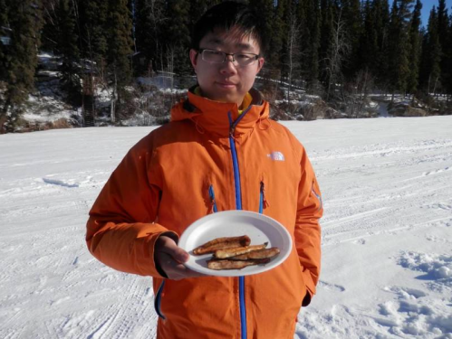 A person in an orange jacket holds a plate of food while standing on snow-covered ground with trees in the background.