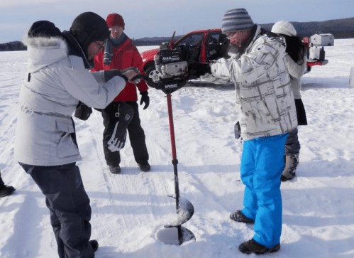 A group of people drills a hole in the snow, with a red vehicle in the background on a snowy landscape.