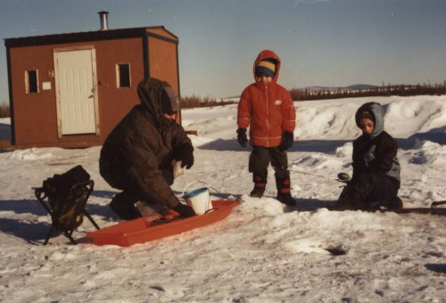 A person and two children in winter clothing ice fishing on a snowy landscape, with a small building in the background.