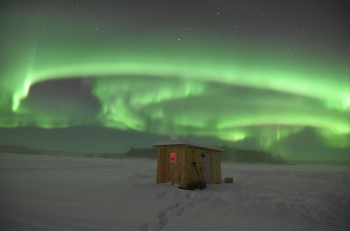 A wooden hut on a snowy landscape under a vibrant green aurora borealis in the night sky.