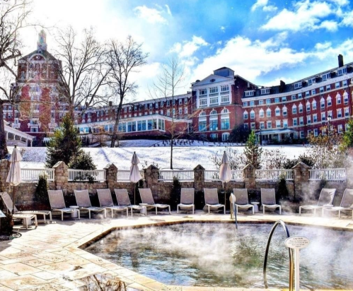 A steaming hot tub surrounded by lounge chairs, with a historic hotel and snowy landscape in the background.