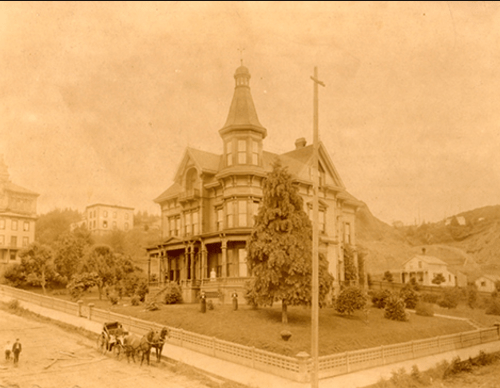 Historic sepia-toned photo of a large Victorian house with a horse-drawn carriage in front and trees surrounding the property.