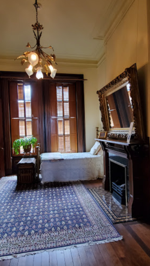 A cozy room featuring a bed, ornate mirror, chandelier, and a patterned rug, with wooden shutters and plants.