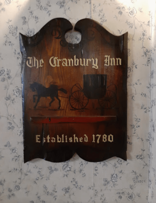 Sign for "The Cranbury Inn," featuring a horse and carriage design, established in 1780, mounted on a floral wallpaper background.
