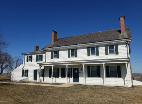 A two-story white house with green shutters and a porch, set against a clear blue sky and grassy landscape.