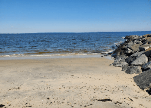 A sandy beach with gentle waves lapping at the shore, bordered by a rocky jetty under a clear blue sky.