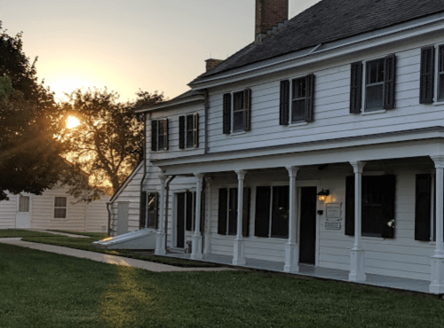 A white house with a porch, set against a sunset, surrounded by green grass and trees.
