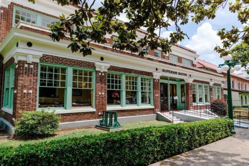 A brick building with green accents, featuring large windows and a sign that reads "Superior Baths," surrounded by greenery.