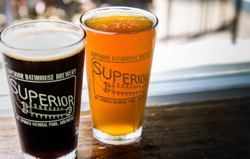 Two glasses of beer, one dark and one light, with "Superior Bathhouse Brewery" logo, set on a wooden table.