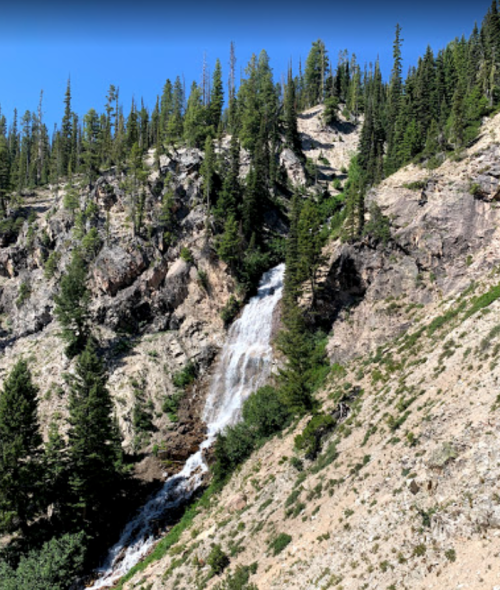 A waterfall cascading down a rocky hillside, surrounded by lush green trees under a clear blue sky.