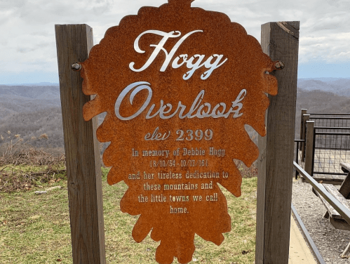 Rustic metal sign reading "Hogg Overlook" in memory of Debbie Hogg, with a scenic mountain view in the background.
