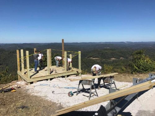 Workers constructing a wooden structure on a hillside with a scenic mountain view in the background.