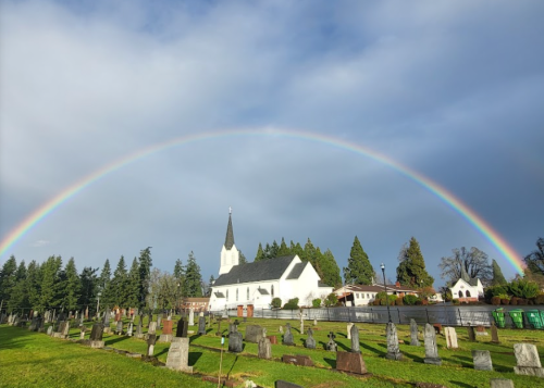 A rainbow arches over a white church and a cemetery, surrounded by trees under a partly cloudy sky.