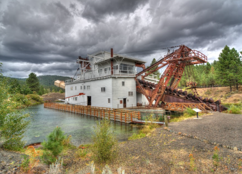 Historic mining structure by a river, surrounded by trees and under a cloudy sky.