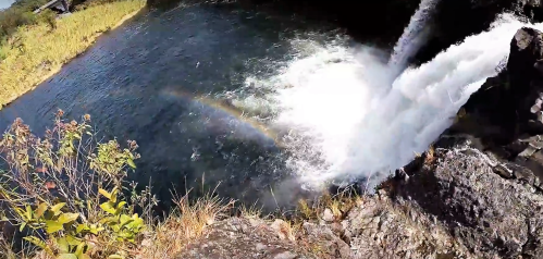 A waterfall cascades into a river, creating a mist with a faint rainbow visible in the spray. Lush greenery surrounds the scene.