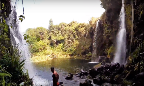 A person stands near a serene waterfall surrounded by lush greenery and rocky terrain.
