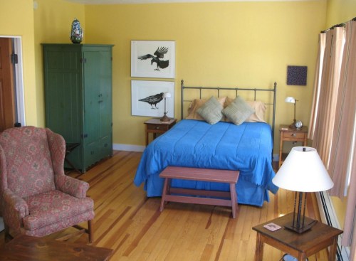 A cozy bedroom with a blue bedspread, wooden furniture, and yellow walls, featuring bird artwork and a patterned chair.
