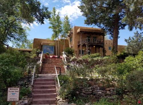 A charming adobe inn surrounded by greenery, featuring a welcoming sign and a staircase leading to the entrance.