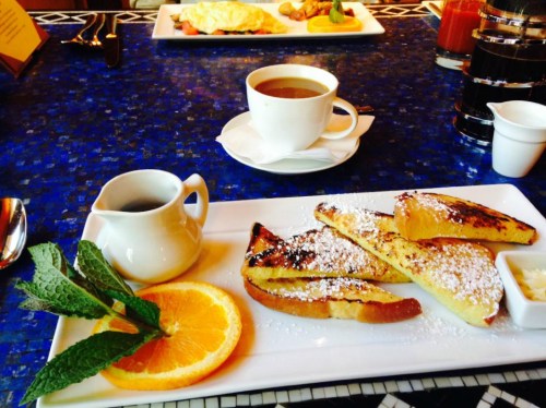A plate of French toast dusted with powdered sugar, orange slices, mint leaves, and a cup of coffee on a blue table.