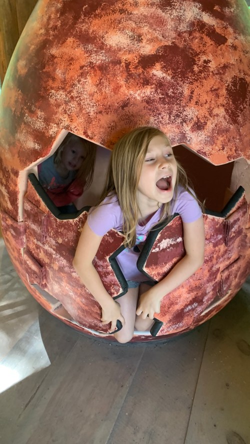 Two children play inside a large, cracked egg structure; one is laughing while the other peeks out from behind.