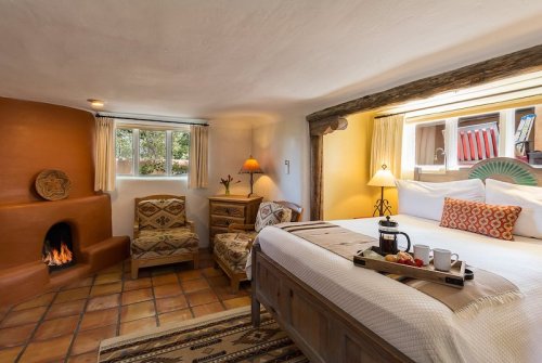 Cozy hotel room with a bed, armchair, fireplace, and a tray of breakfast on the bed. Warm, inviting decor.