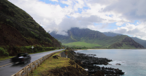 A scenic coastal road winds along rocky shores, with lush mountains and clouds in the background.