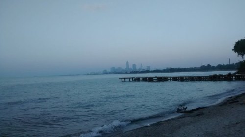 A serene lakeshore view with a distant city skyline under a hazy sky, reflecting calm waters and a sandy beach.