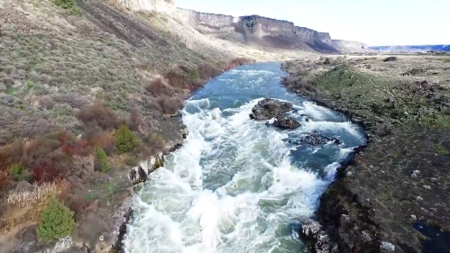 Aerial view of a rushing river surrounded by rocky banks and grassy hills under a clear blue sky.