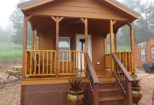 A cozy wooden cabin with a porch, steps, and decorative pots, surrounded by greenery in a misty setting.