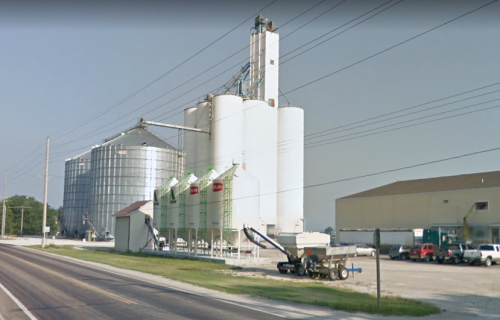 Grain silos and storage facilities alongside a road, with machinery and power lines in the foreground.