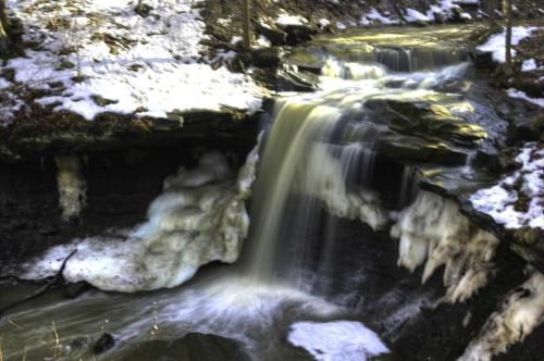 A serene waterfall cascades over rocks, surrounded by snow and ice in a tranquil forest setting.