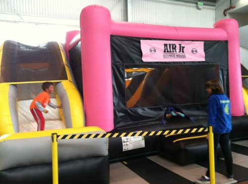 A colorful bounce house with a slide, featuring children playing and an adult supervising in a spacious indoor area.
