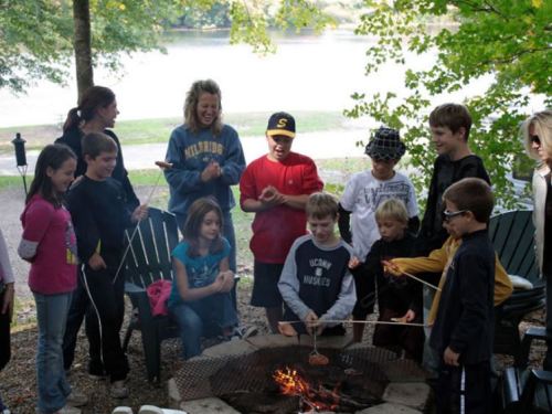 A group of children and adults gather around a campfire, roasting marshmallows and enjoying a sunny day outdoors.