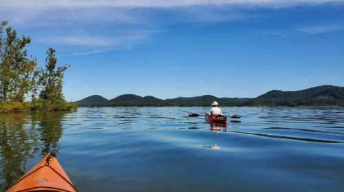 A person in a kayak paddles on calm water, surrounded by trees and distant hills under a clear blue sky.