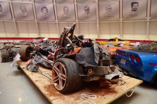 A damaged car wreck on a display platform, surrounded by other vehicles in a museum-like setting.