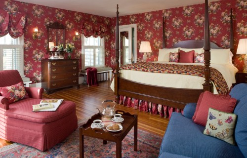 Cozy bedroom with floral wallpaper, a four-poster bed, seating area, and a tea set on a table. Warm, inviting decor.