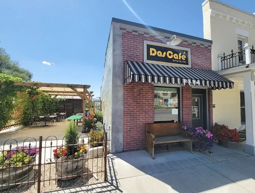 A brick café building with a striped awning, surrounded by colorful flowers and a patio area with seating.