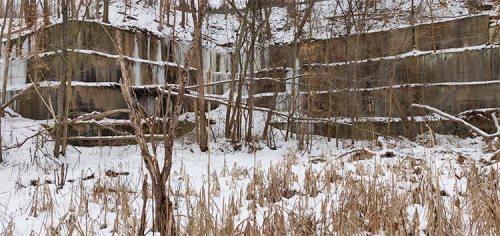Abandoned concrete structures partially covered in snow, surrounded by trees and tall grass in a winter landscape.