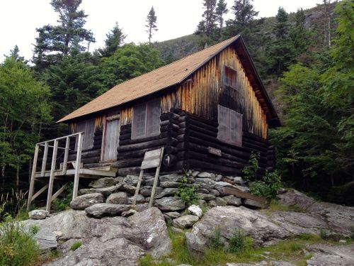 A rustic log cabin on rocky terrain, surrounded by trees and mountains, with a wooden porch and weathered sign.