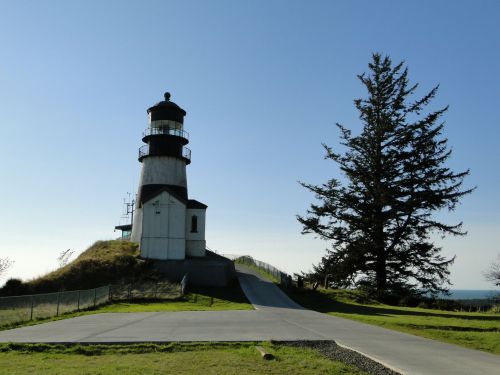 A tall lighthouse stands on a grassy hill, with a large tree nearby and a clear blue sky in the background.