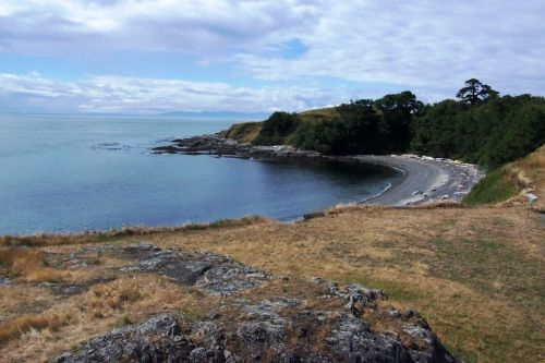 A serene coastal view with a curved beach, rocky shoreline, and lush greenery under a partly cloudy sky.