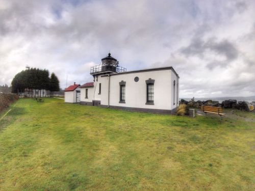 A white lighthouse with a black lantern room, surrounded by green grass and cloudy skies.
