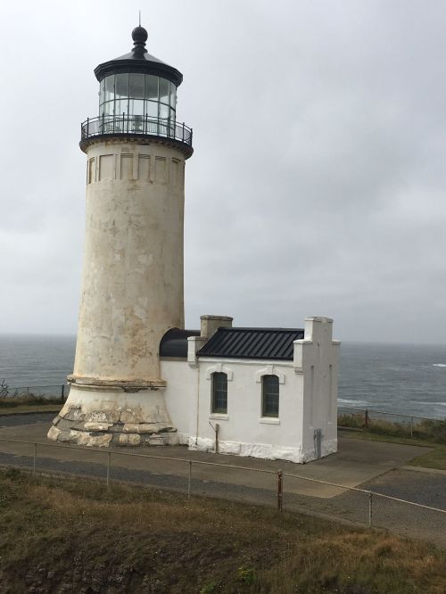 A tall lighthouse stands beside the ocean, with a white building at its base, under a cloudy sky.