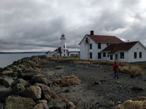 A person walks along a rocky path near a lighthouse and a white house under a cloudy sky by the water.