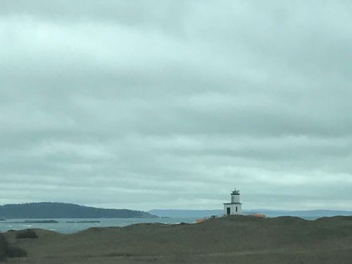 A small white lighthouse stands on a grassy shore, with a cloudy sky and distant hills in the background.