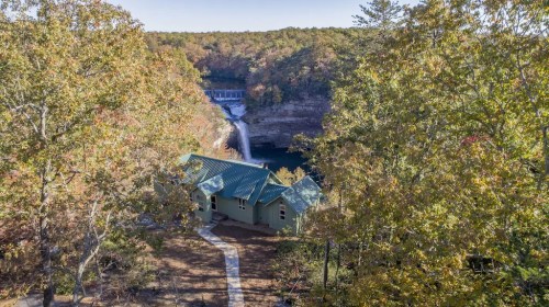 Aerial view of a green house surrounded by autumn trees, overlooking a waterfall and a serene body of water.