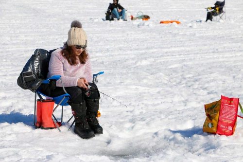 A person sits on a chair in the snow, ice fishing with a rod, surrounded by a snowy landscape and other fishers.