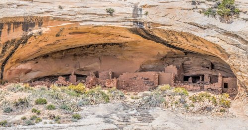 Ancient stone ruins nestled under a large rock overhang, surrounded by desert vegetation.